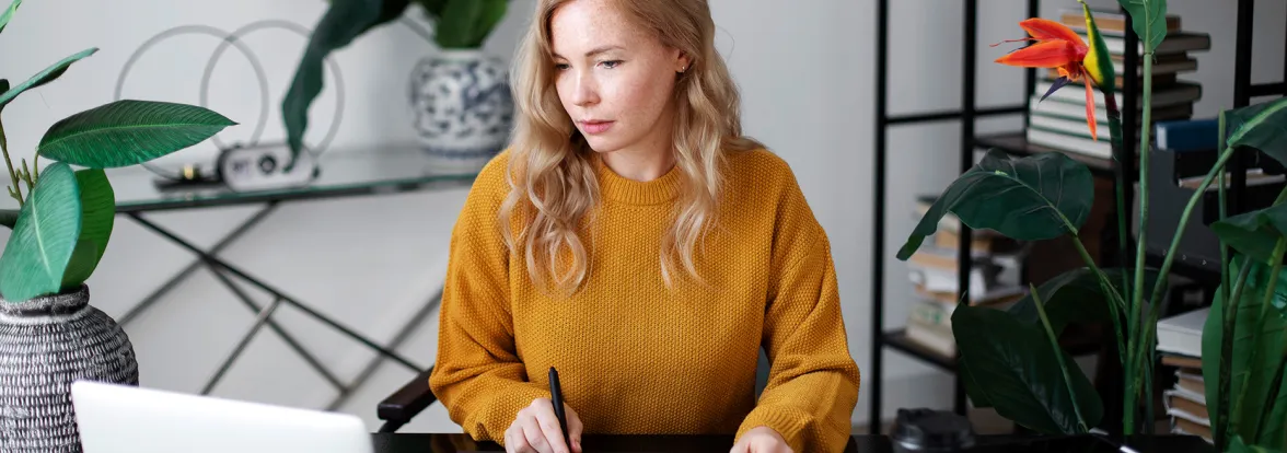 A woman in a white woman holds a pen in one hand while smiling at her laptop