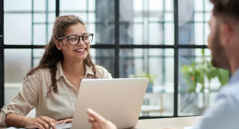 A woman smiles at a colleague and looks up from her laptop