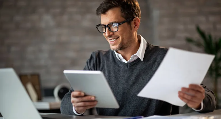 A man in business casual holds papers in his hands at a work desk