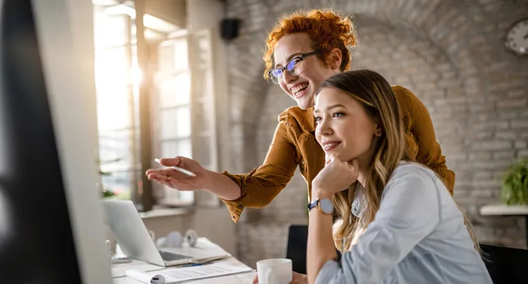 A woman standing looks over her colleague to point at something on her computer