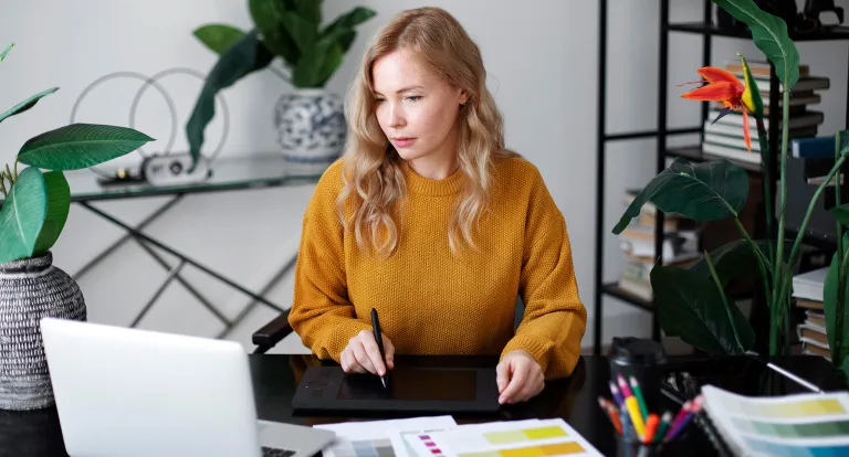 A woman at a desk holds a pen surrounded by papers and a laptop