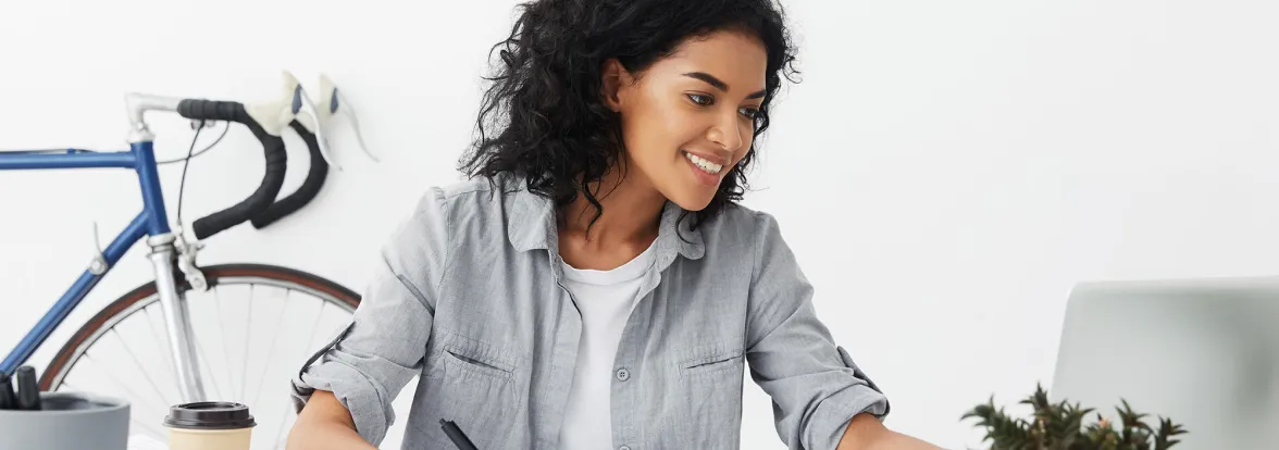 A woman in a white woman holds a pen in one hand while smiling at her laptop