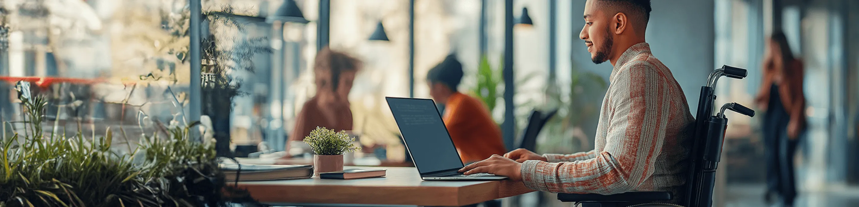 A man in a wheelchair sits at his laptop in a café with other people in the background