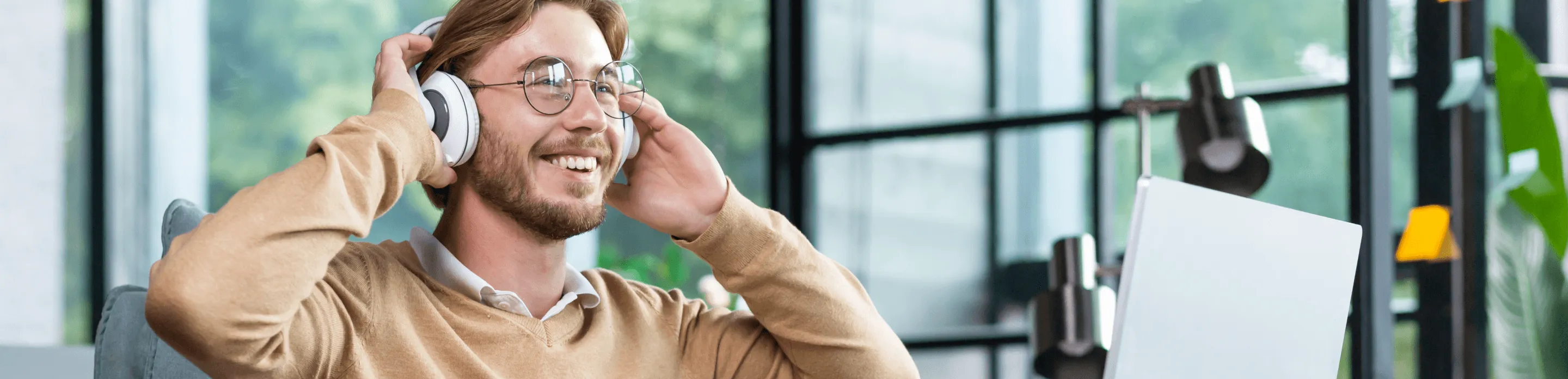 A man wearing glasses smiles as he touches his headphones and looks up from laptop