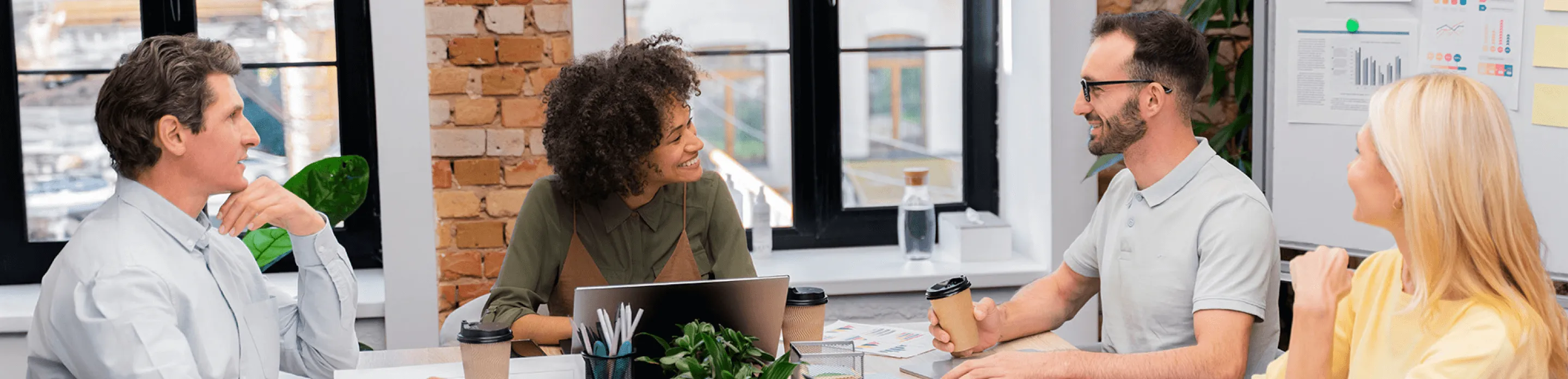 Four colleagues smile in a meeting room with coffees and graphs on whiteboards behind them