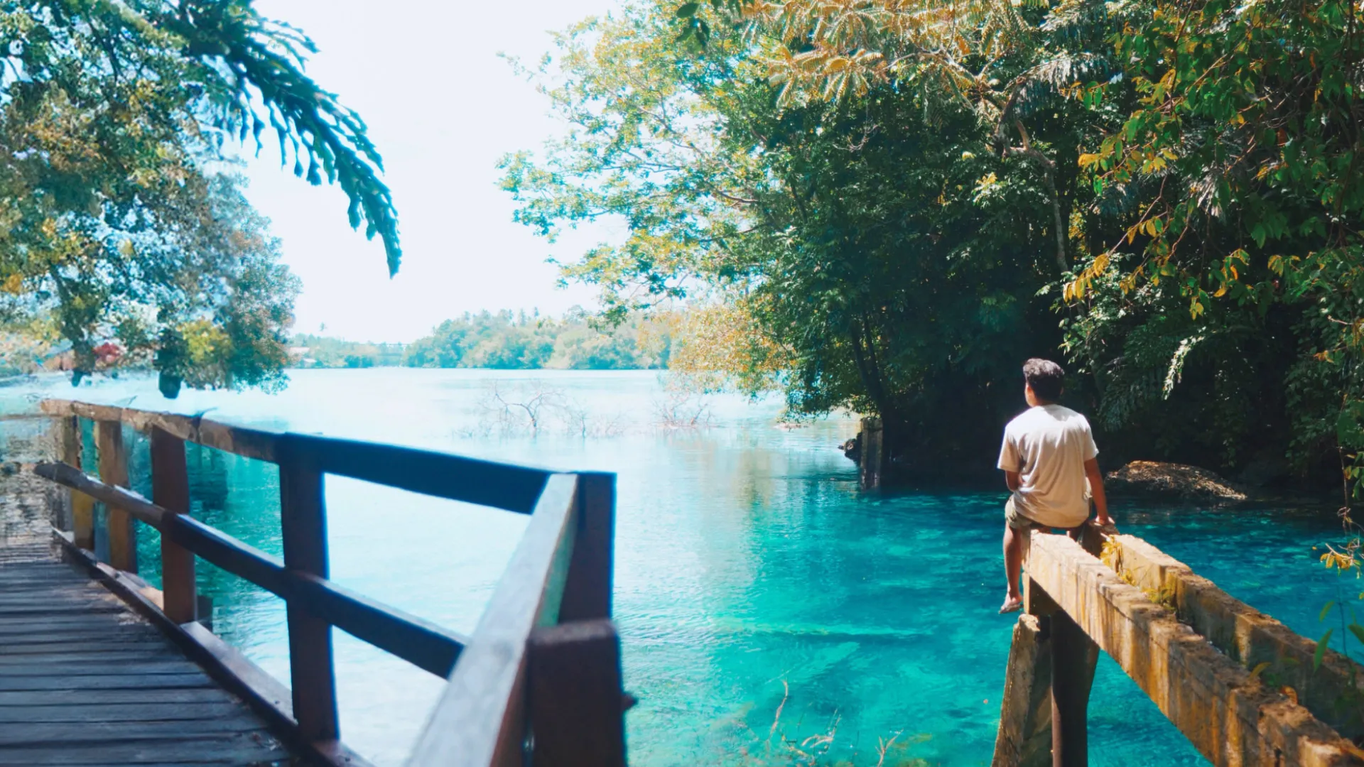 A man in shorts and t-shirt sits at the end of a wooden structure above glistening turquoise water