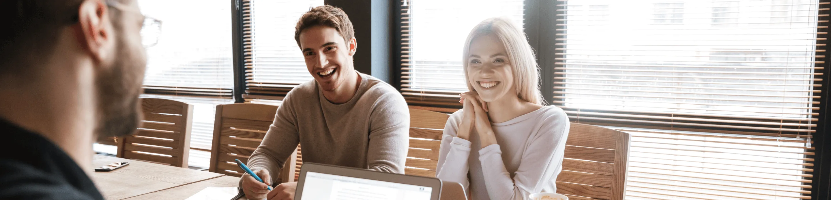 Three people smile and collaborate across a wooden table with laptops and pens