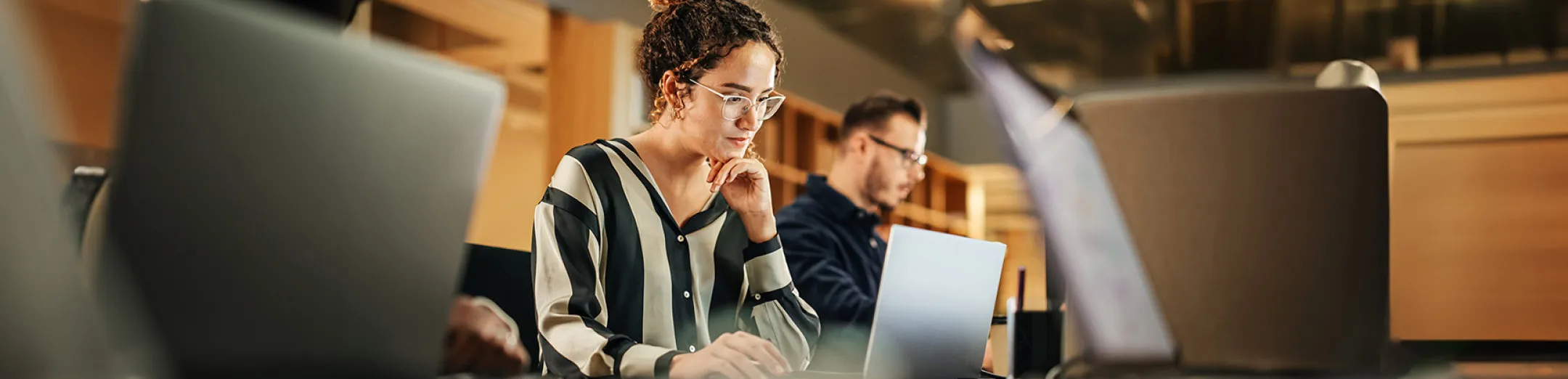 Woman looks intently at her laptop screen at a coworking space, with an out of focus man in the background