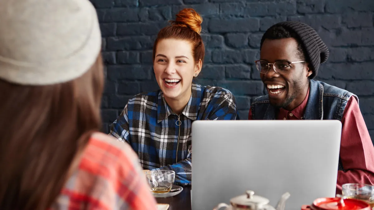 Three people sit and laugh around a laptop at a café