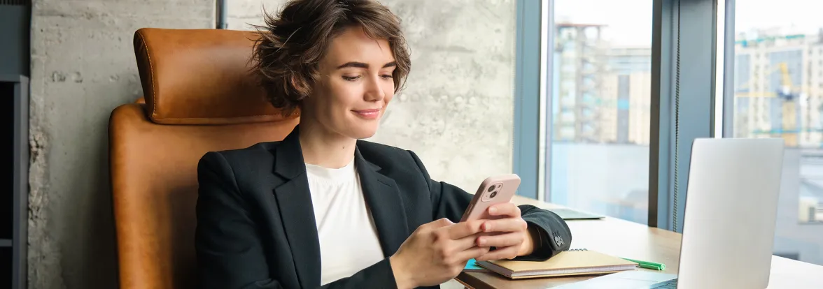 A seated woman in a suit jacket looks at her phone as she leans on her desk